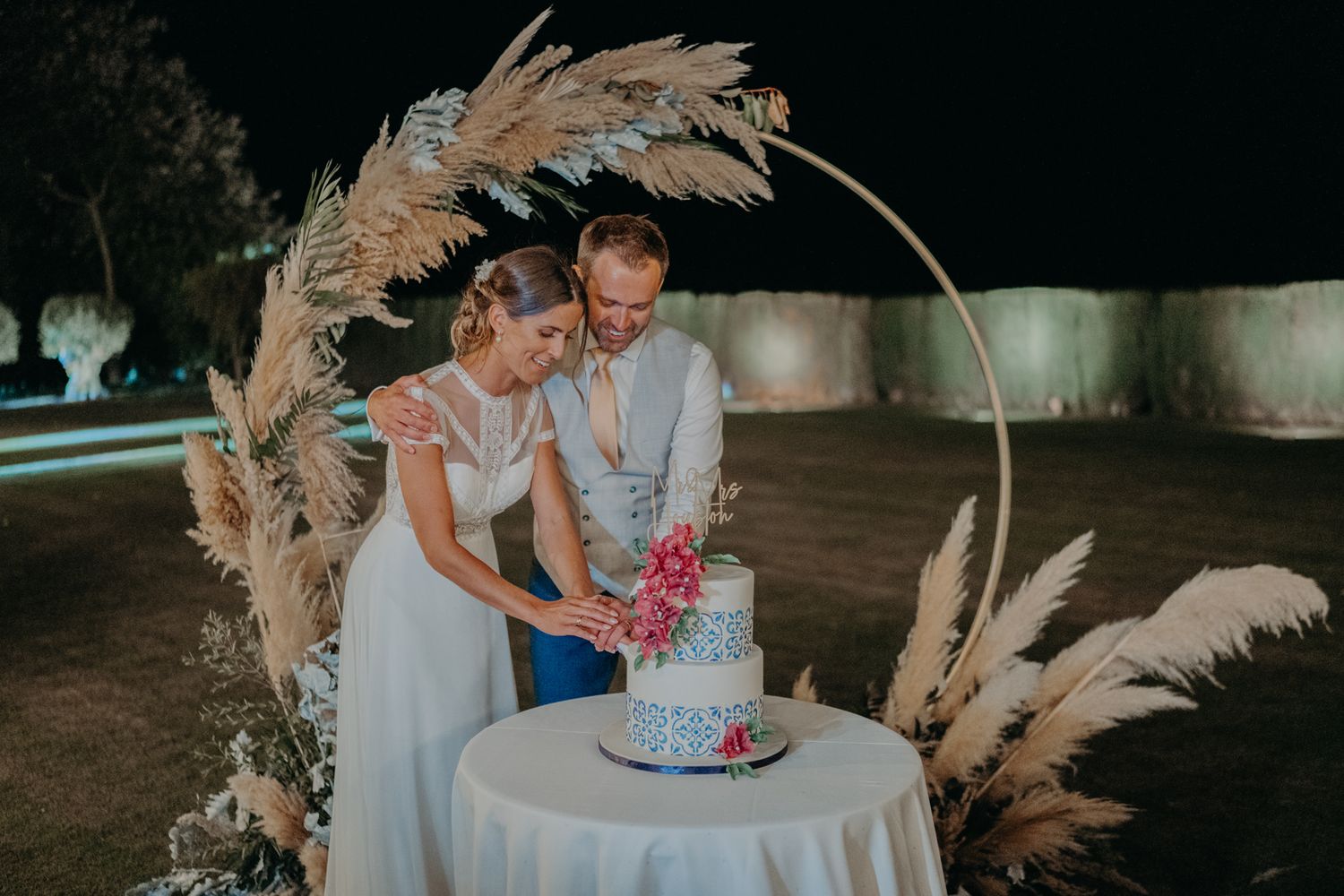 A wedding cake cutting moment captured beneath a curved arch adorned with dried pampas grass.
