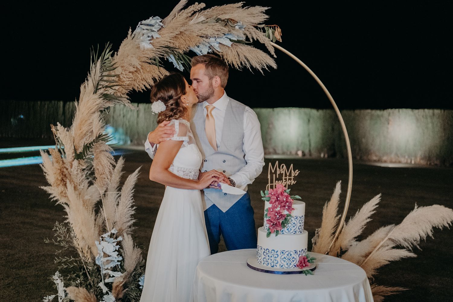 A couple celebrates their wedding beside a circular arch decorated with pampas grass and a tiered blue and pink cake.