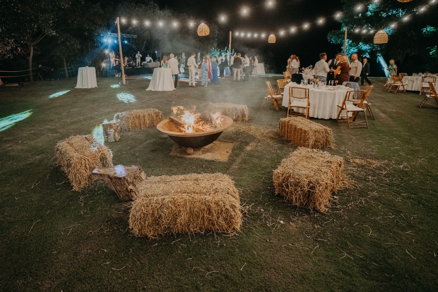 Rustic outdoor wedding reception setup with hay bales around a fire pit and string lights overhead.