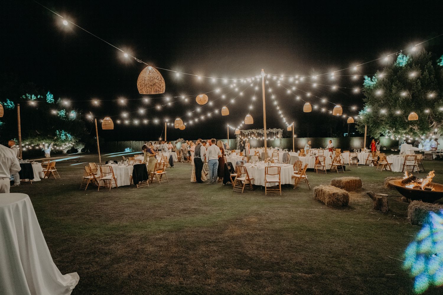 String lights create a magical atmosphere over an outdoor wedding reception setup with scattered dining tables.