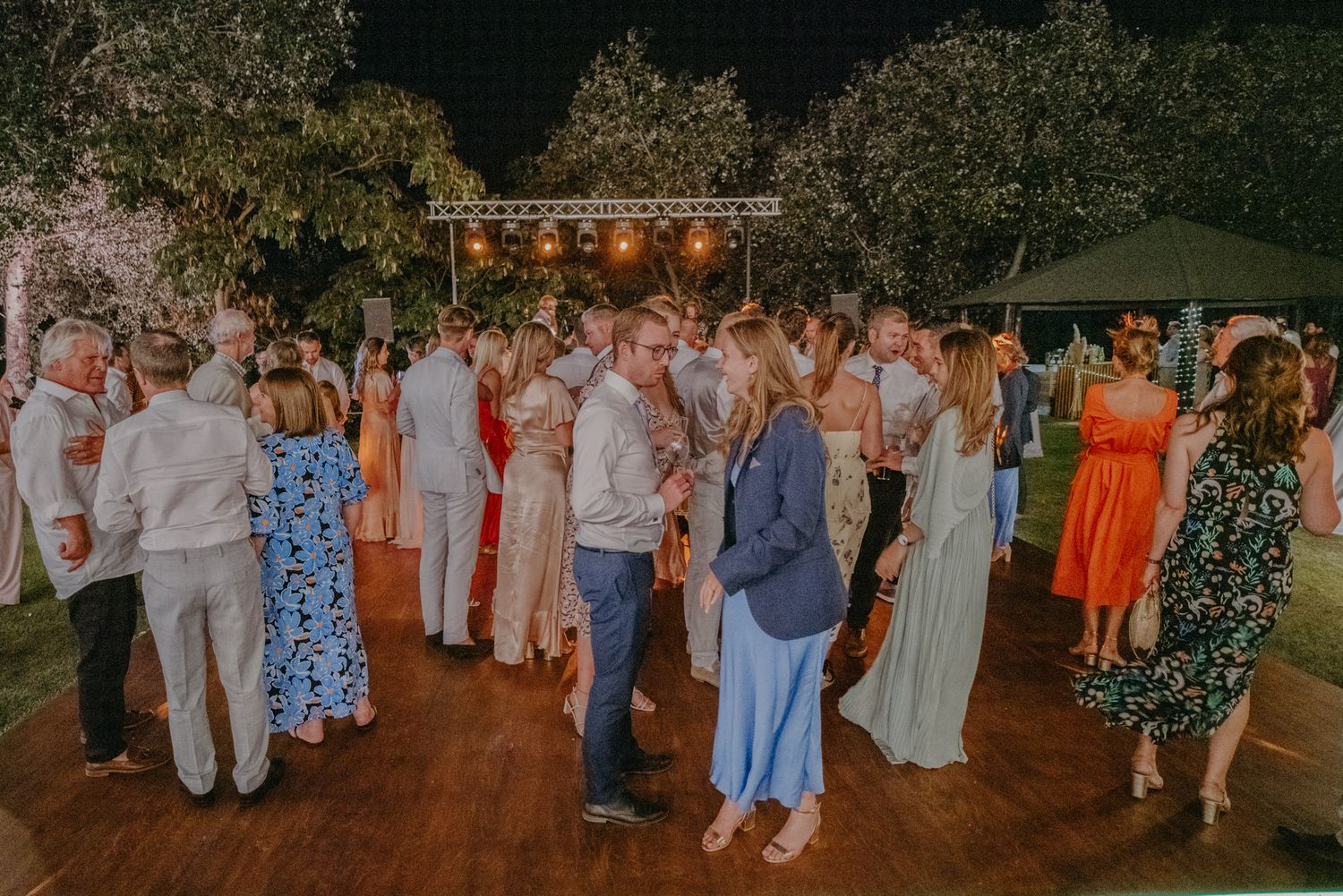 Wedding guests dance on an outdoor dance floor under string lights at dusk during a festive reception celebration.