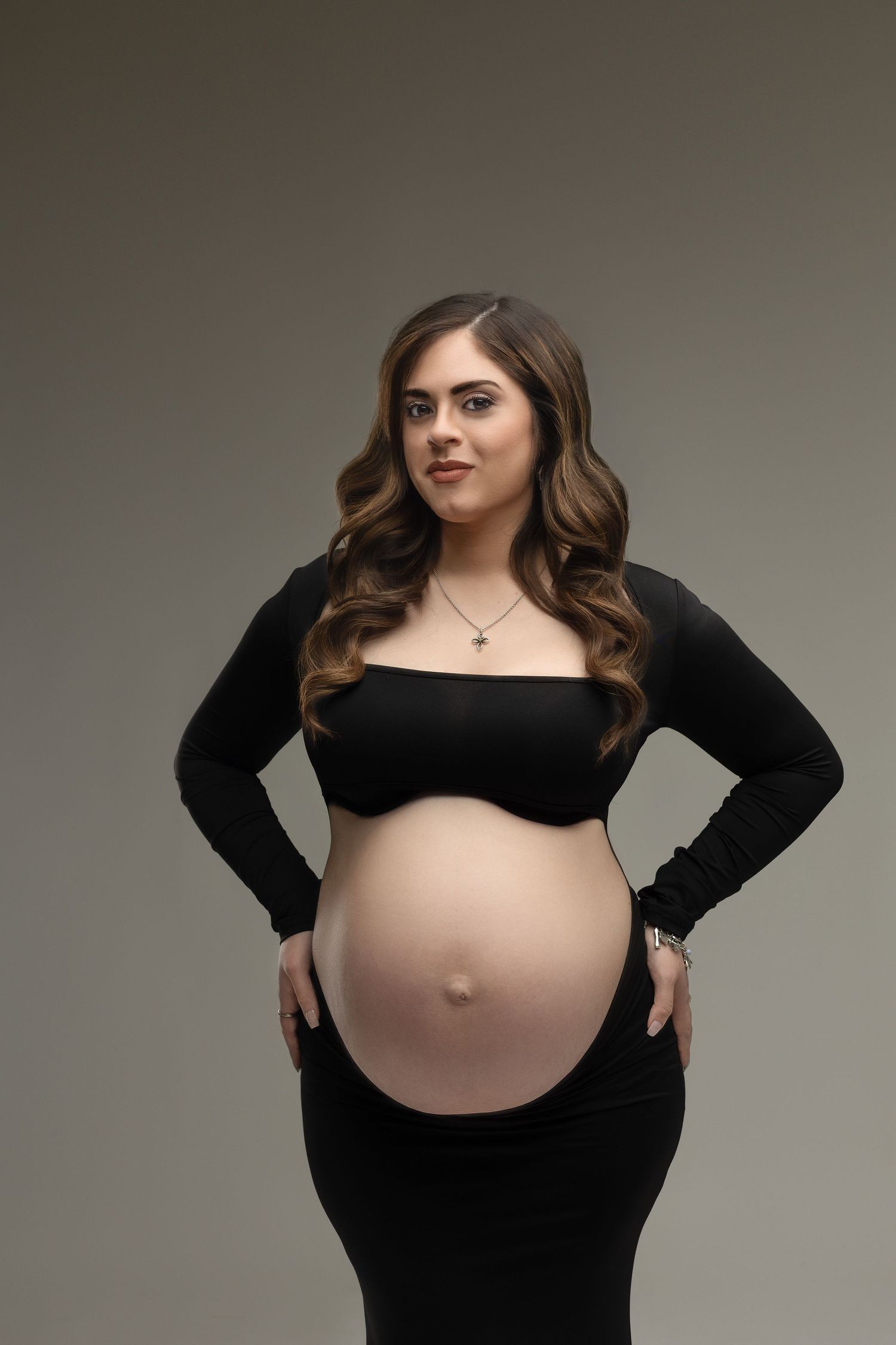 Expectant mother wearing black dress poses against gray background in elegant maternity photoshoot.