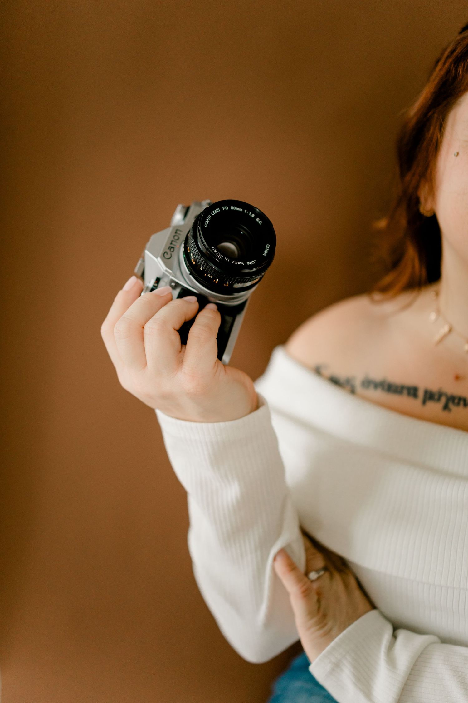 Woman in white off-shoulder top holding a vintage film camera with tattoo visible on chest.