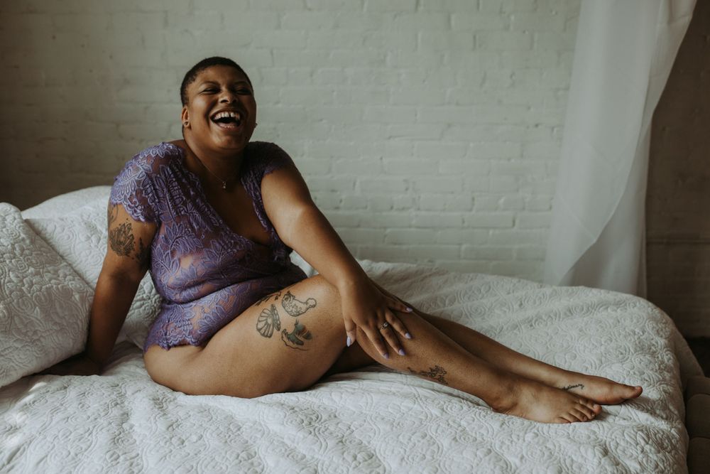 A joyful figure wearing purple loungewear relaxes on a white bed against a brick wall background.