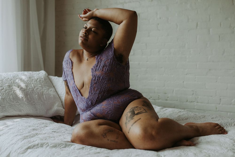 A person in a purple lingerie poses artistically on a white bed against a brick wall.
