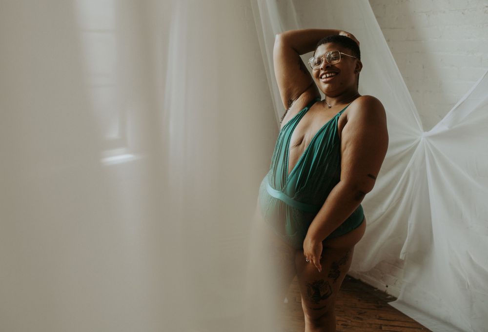 Person in emerald green plunging swimsuit poses gracefully against white draped backdrop in moody boudoir setting.
