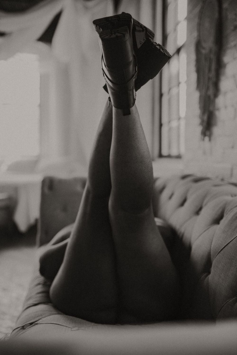 Black and white photo of crossed legs in knee-high boots resting on a plush couch in moody lighting.