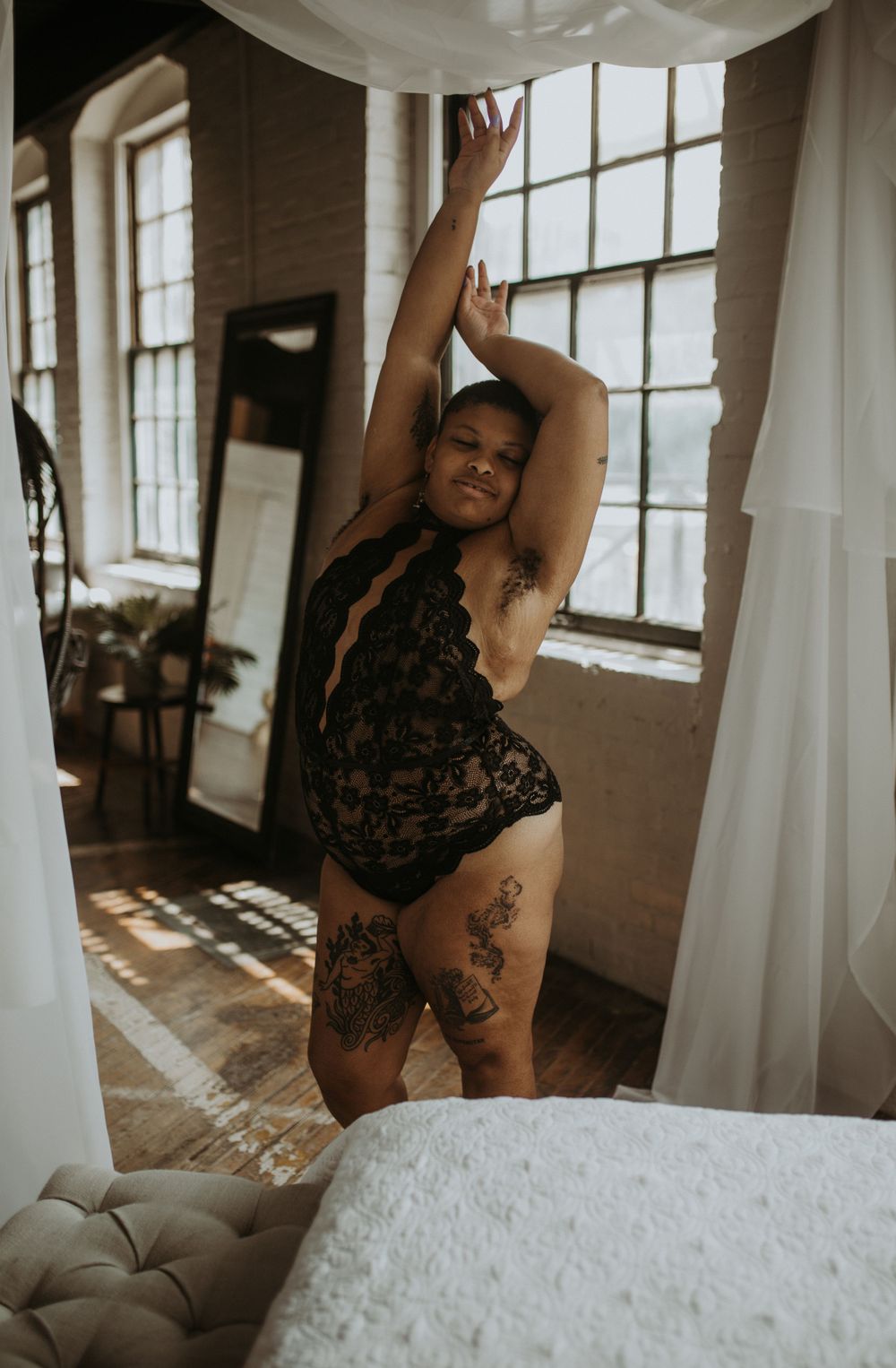 A person in black lace lingerie poses by a sunlit window displaying natural confidence and body positivity.