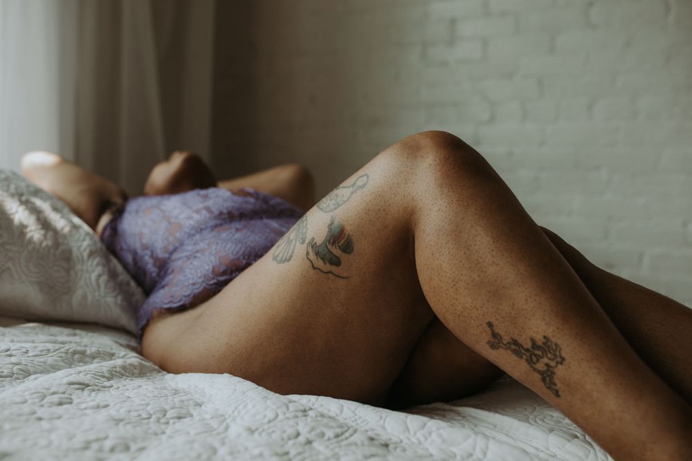 Tattoos adorn bare legs resting on white bedsheets in a moody boudoir setting.