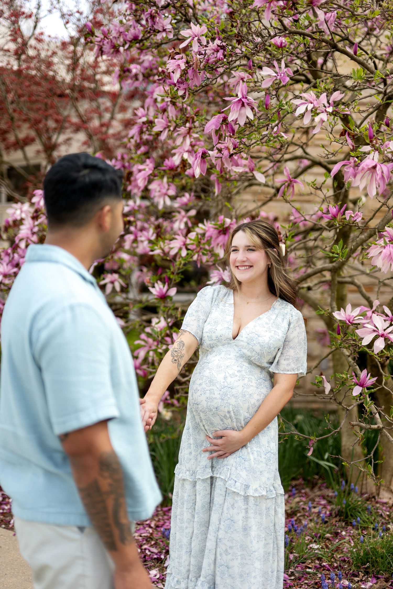 Pregnant woman in floral dress holds belly, smiling at partner near blooming pink magnolia tree outdoors.
