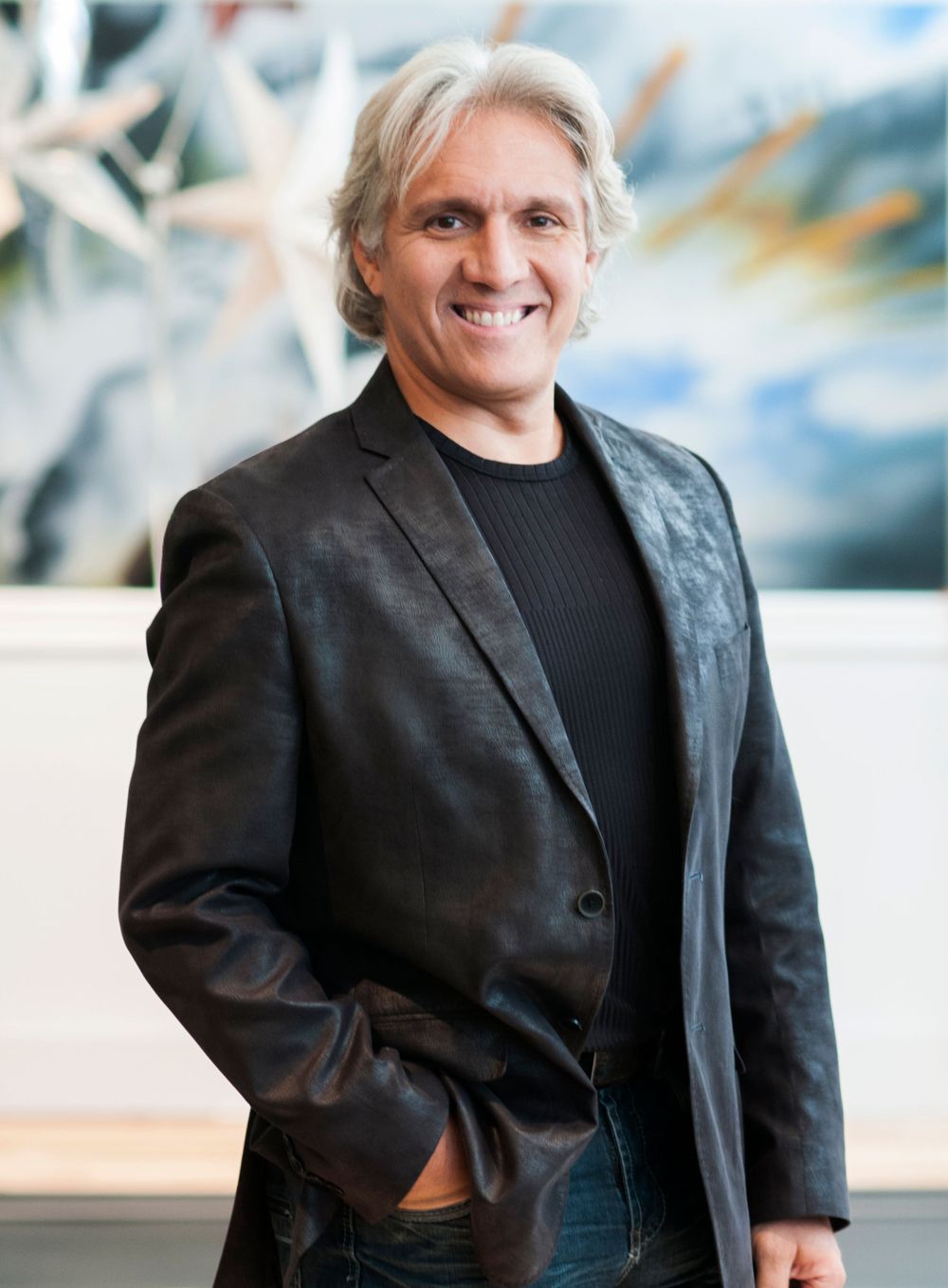 Professional headshot of someone in a black blazer and shirt against an artistic background.