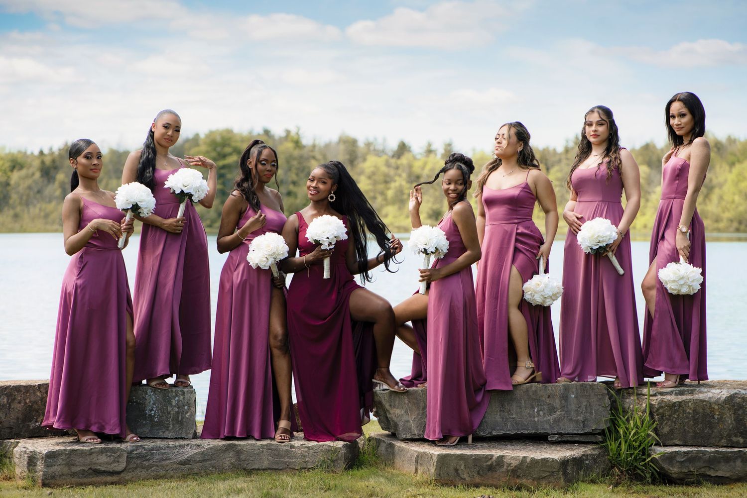 Bridesmaids in flowing mauve gowns holding white floral bouquets pose together on stone steps near a waterfront.