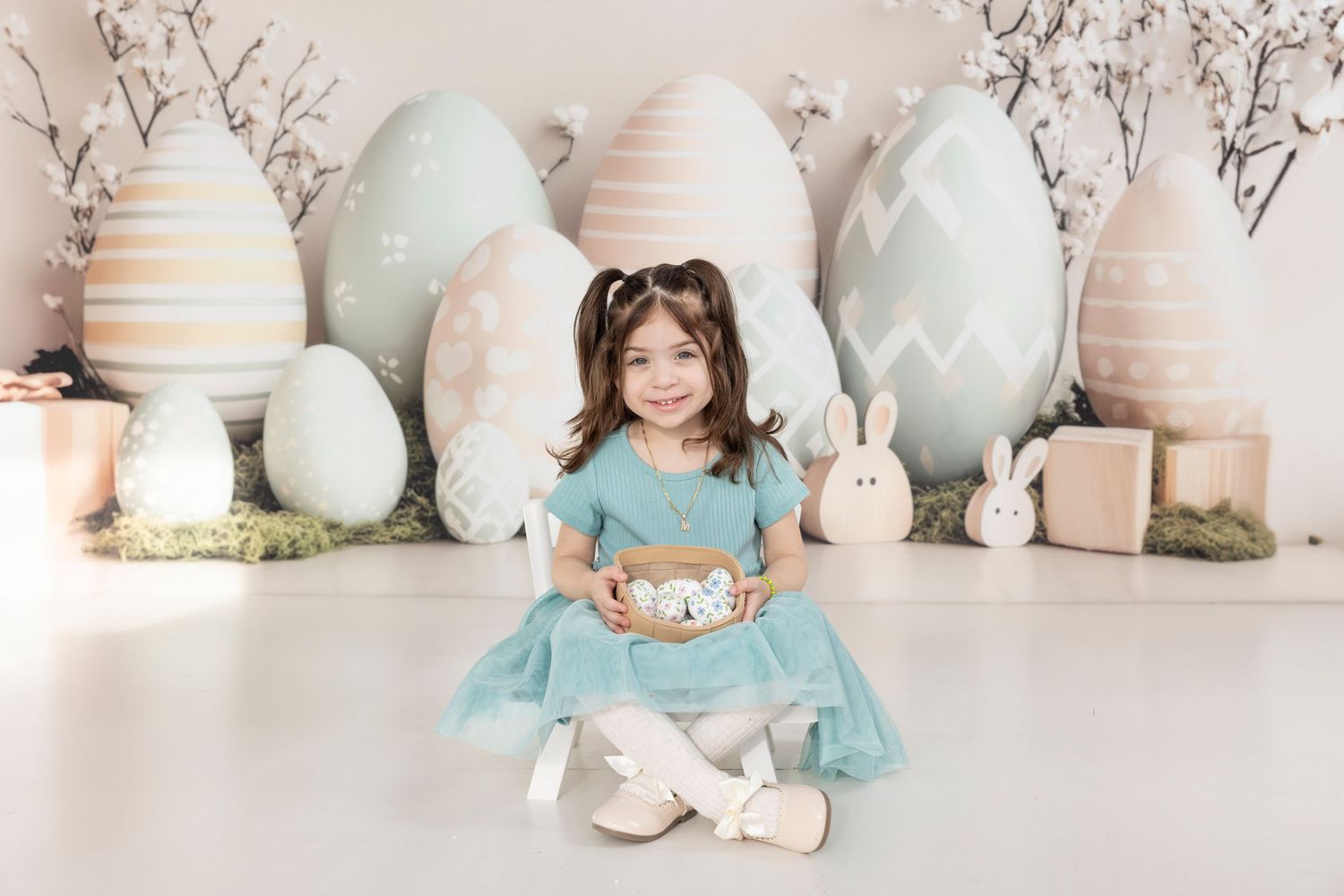 Easter themed portrait setup with large pastel eggs, white bunnies, and a child in turquoise dress sitting on white floor.