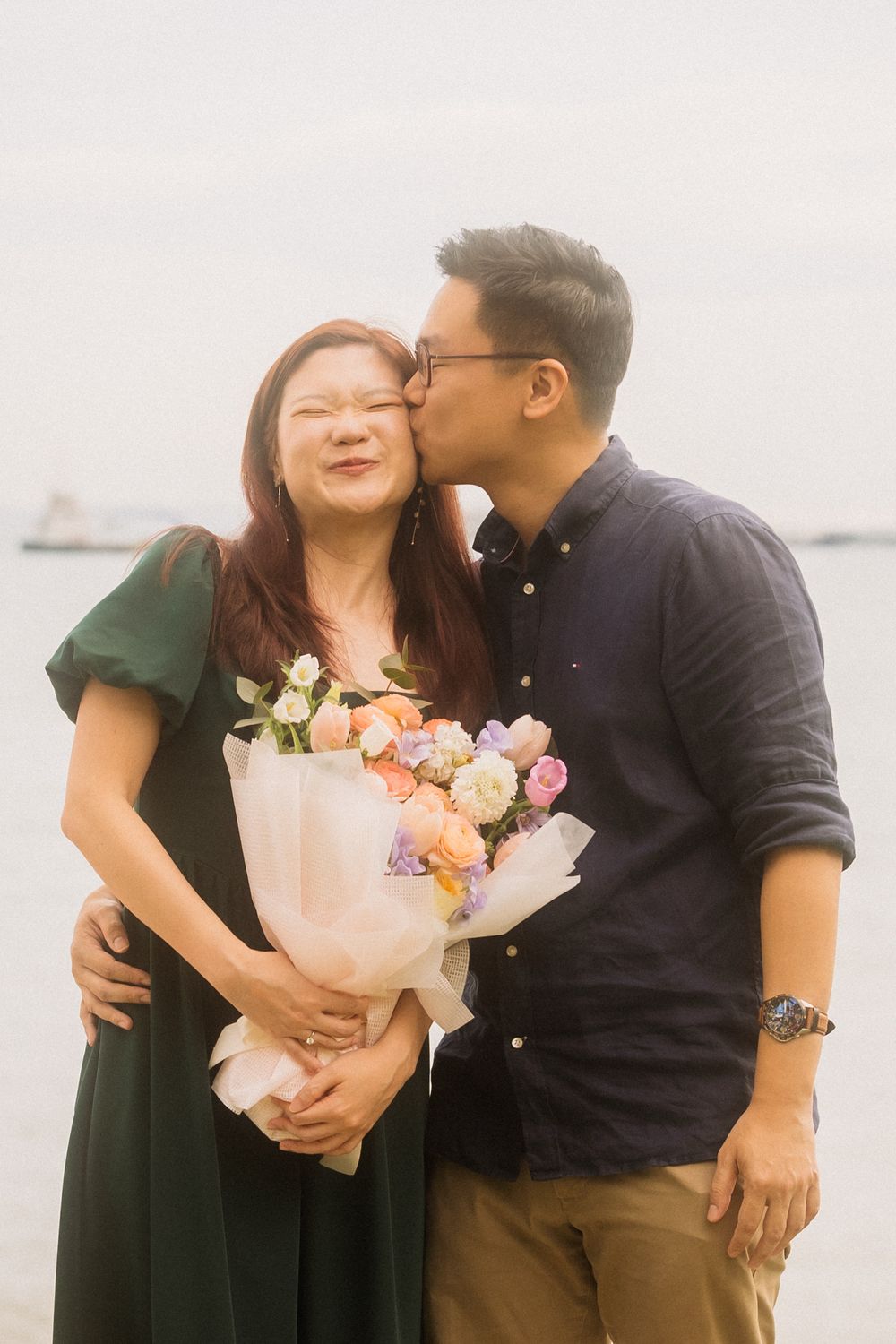 A loving couple shares a tender moment by the water while holding a beautiful bouquet of pink and peach flowers.