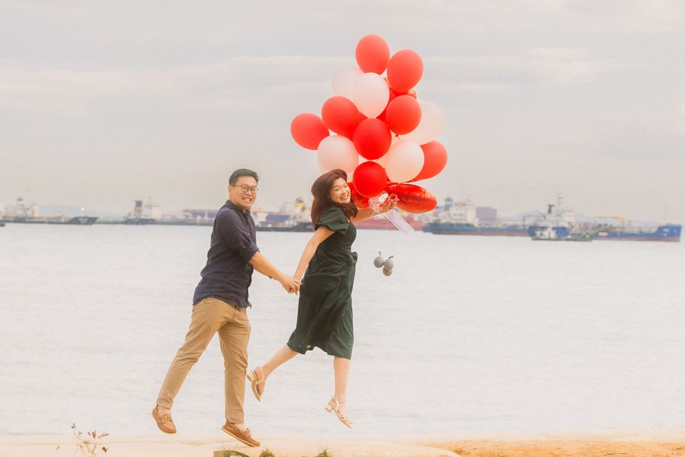 A couple walks along a beach holding red balloons against a cloudy sky with distant ships visible on the horizon.