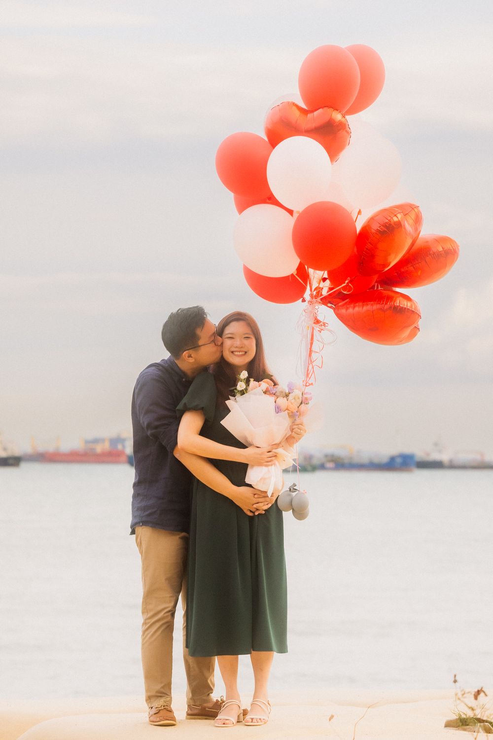 A couple embraces by the water holding red balloons during a romantic waterfront photoshoot at sunset.