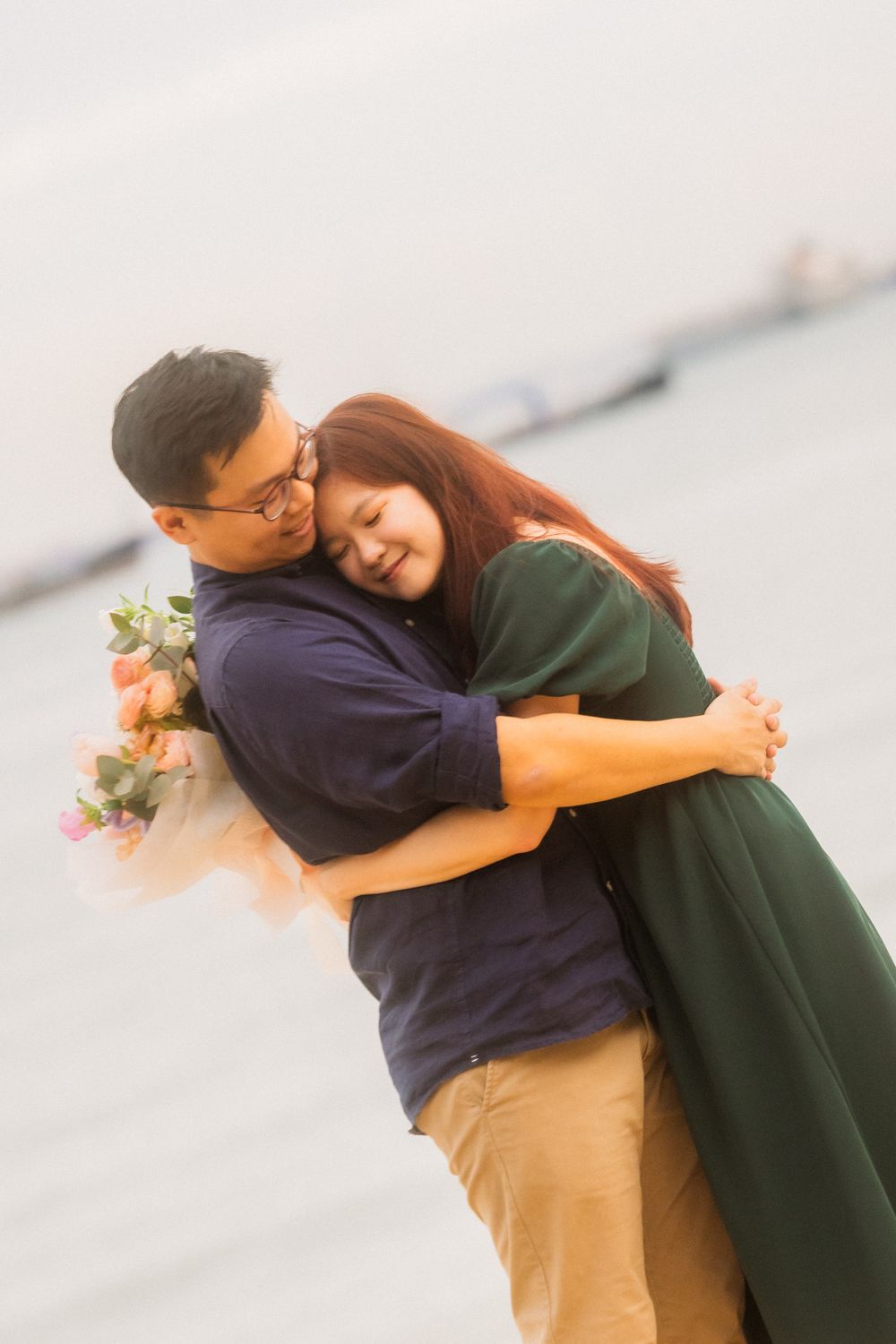 A romantic couple in casual attire sharing an intimate embrace while holding flowers against a soft background.