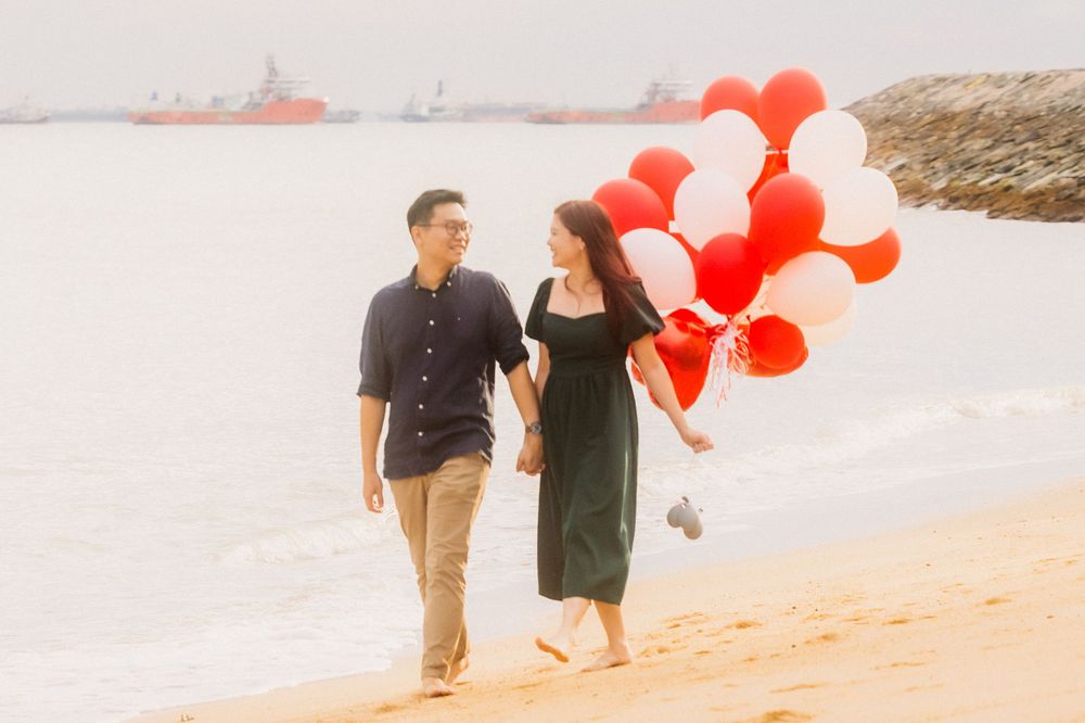 A romantic couple walks along a sandy beach holding red and white balloons during golden hour.