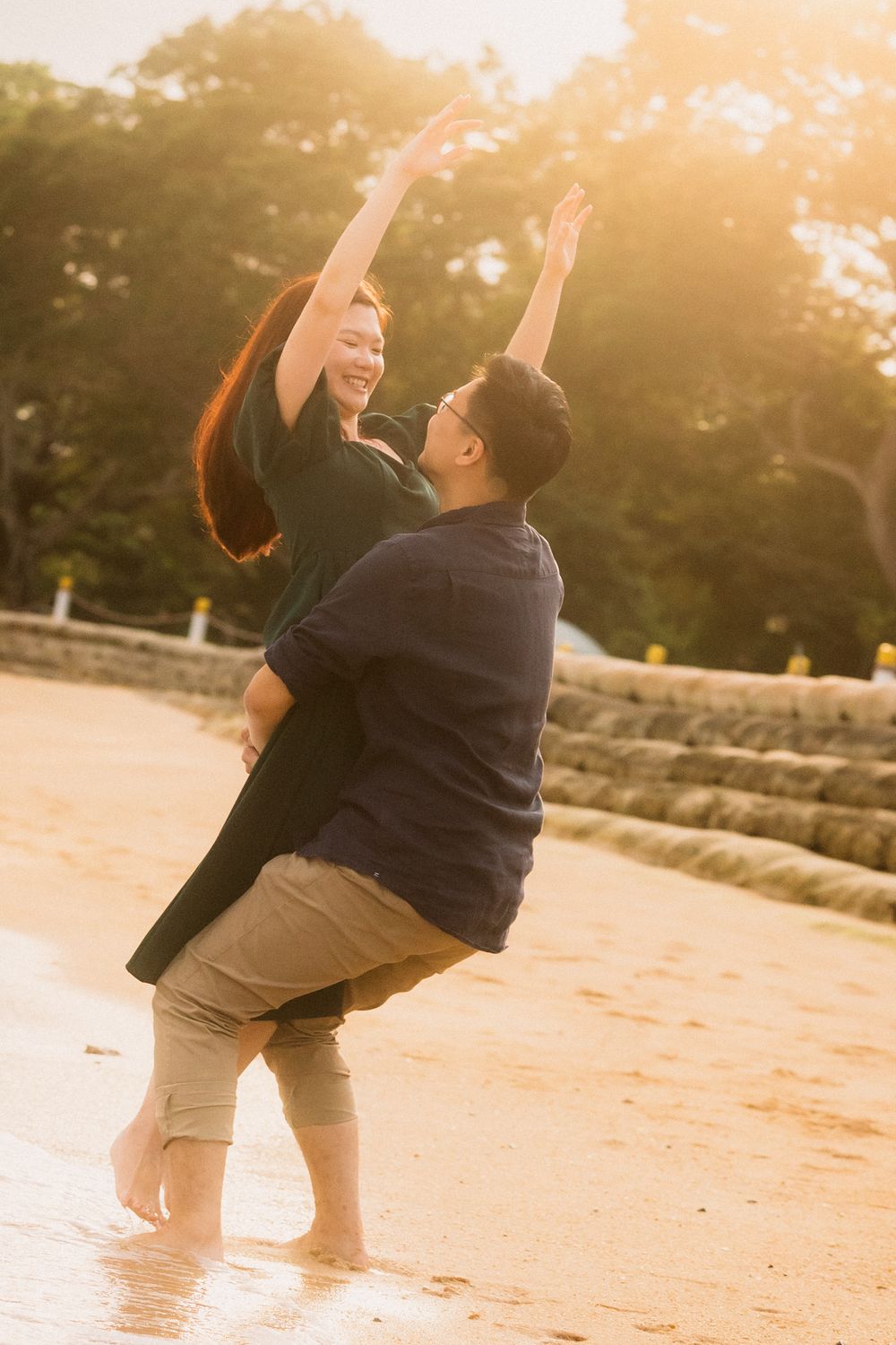 A couple joyfully dances and embraces on a sandy beach at sunset with golden light streaming through the trees.