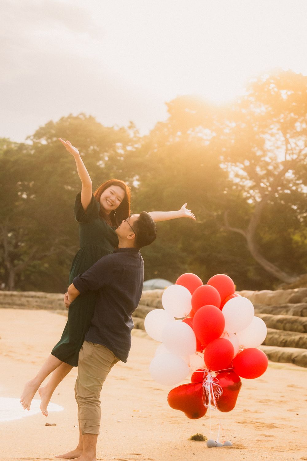 A joyful couple share a playful moment on the beach with red and white balloons at sunset.