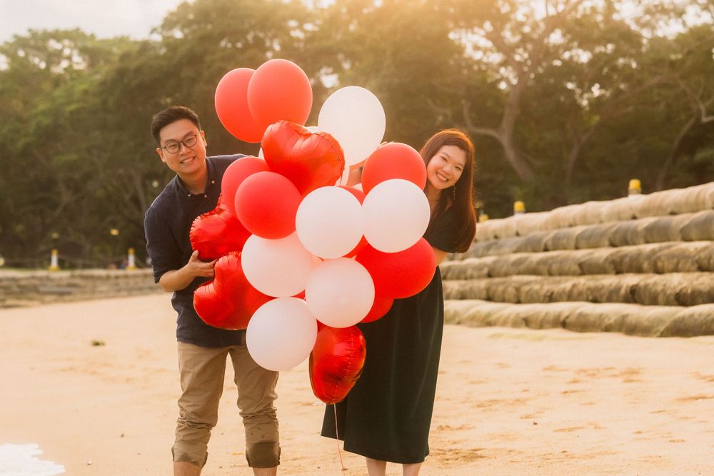 A couple holds red and white balloons during golden hour at a sandy location with stone steps in the background.