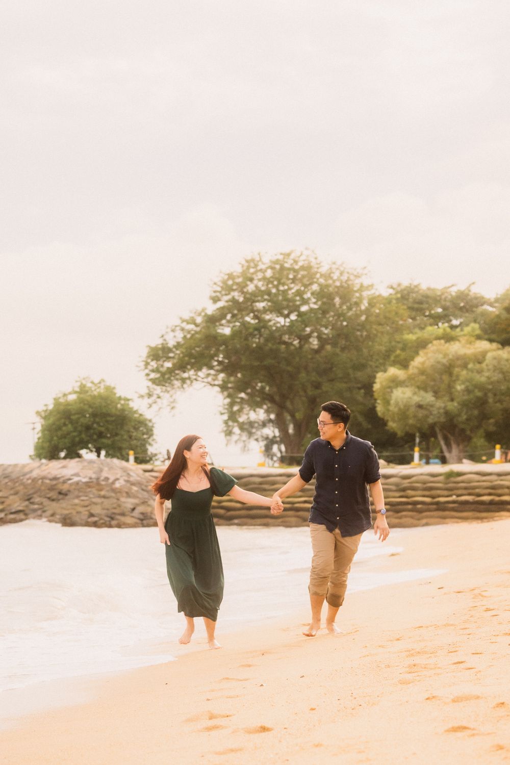 A couple joyfully runs together barefoot along a sandy beach with trees in the background.