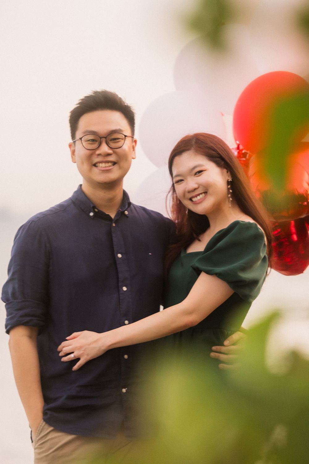A romantic couple poses together for a portrait with red balloons and a soft blurred background.