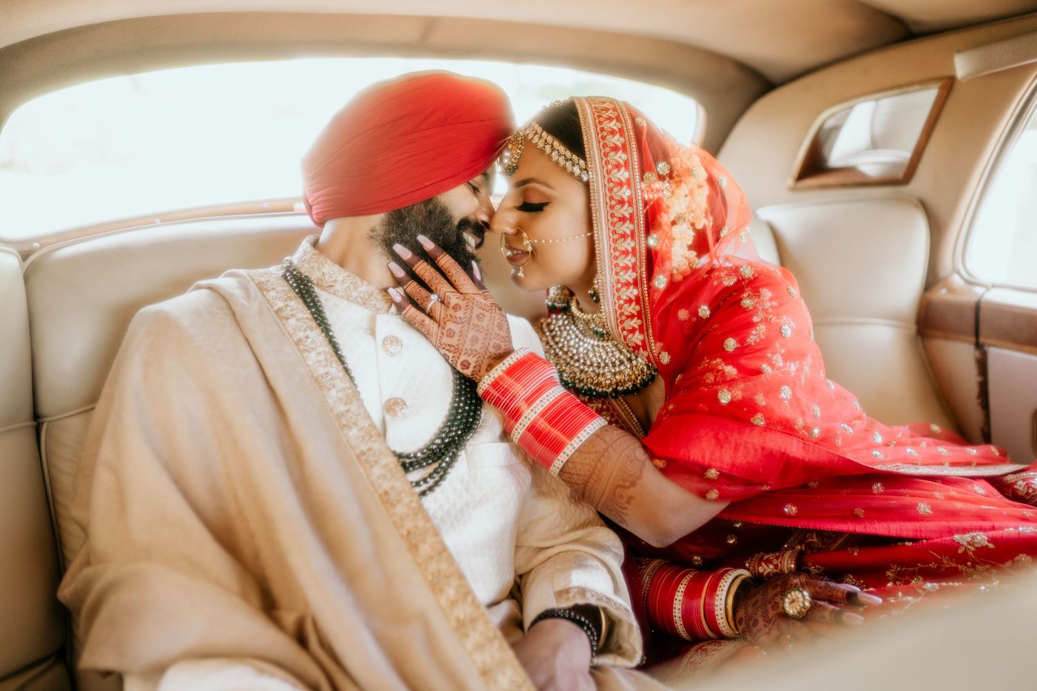Traditional Indian wedding couple shares an intimate moment in the backseat of a car wearing red and cream attire.