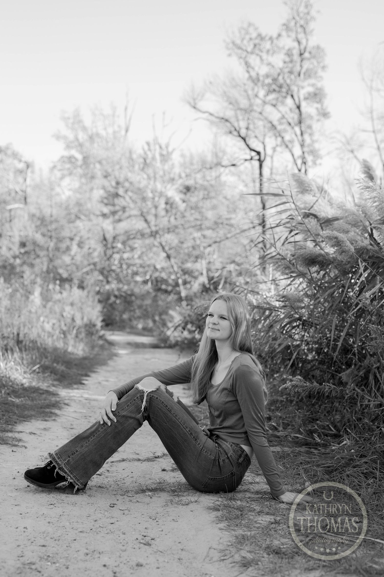 A black and white portrait of someone sitting casually on a dirt path surrounded by trees and foliage.