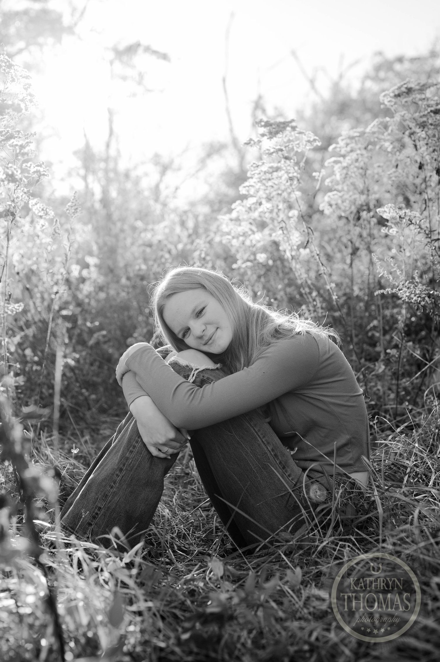 Black and white portrait of person curled up in casual pose among wild grasses.
