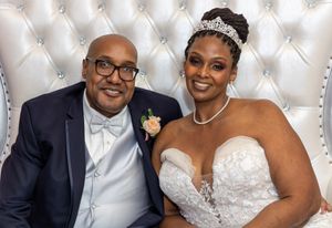 Wedding couple in formal attire pose together against a white tufted backdrop during their celebration.