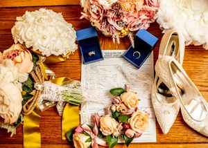 Wedding details with rings, invitation, flowers and bridal shoes laid out on wooden surface.
