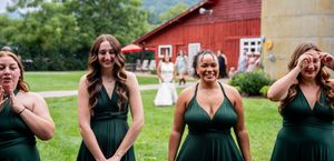 A group in emerald green dresses stands together on grass with a red barn in the background.