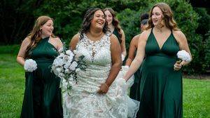 A group of bridesmaids in emerald green dresses and white lace gowns stand together on a grassy lawn.