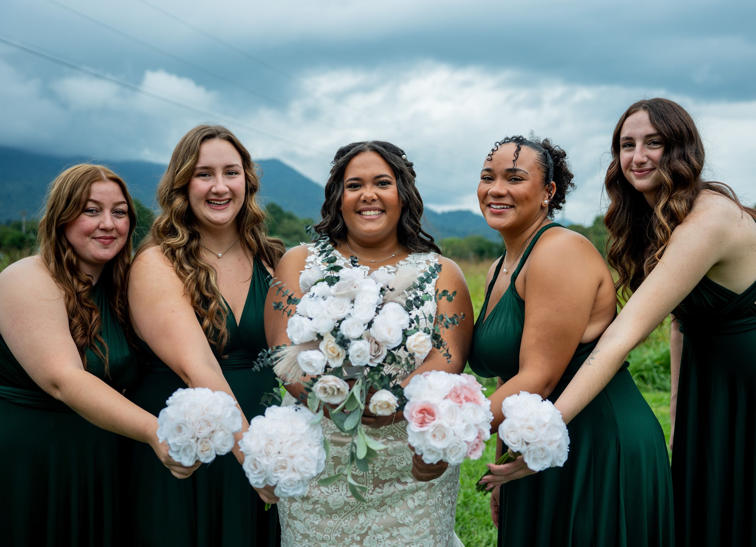 A bridal party in dark green dresses and white bouquets poses together against a mountain landscape backdrop.