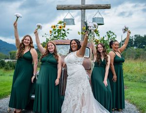 Wedding party in emerald green dresses poses joyfully outdoors with a white wedding gown displayed.