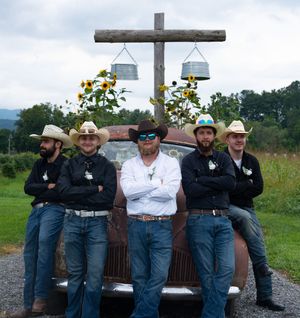 Groomsmen in jeans and dark shirts lean casually against a rustic wooden fence at an outdoor venue.