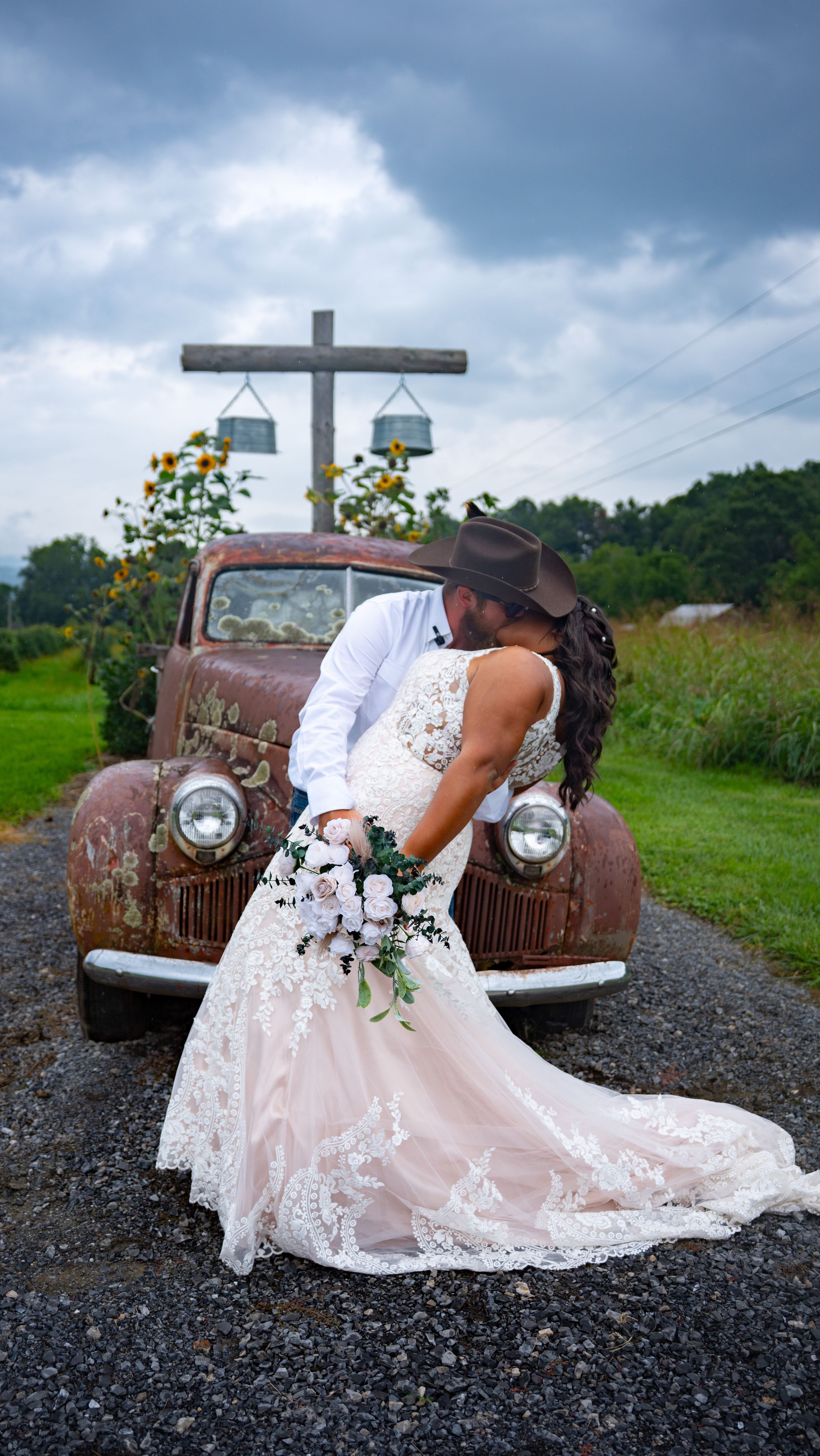 A bride in a lace wedding dress poses dramatically near a vintage rustic truck under stormy skies.