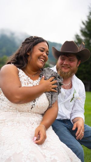 A couple shares joyful laughter during an outdoor photo session, with the bride wearing a white lace dress.