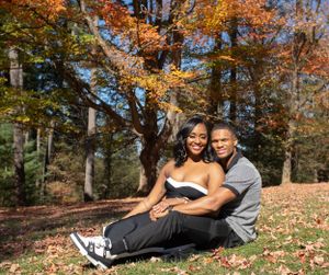 A couple sits together on autumn ground beneath vibrant orange fall foliage in a peaceful forest setting.