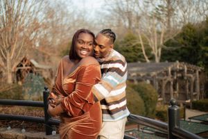 Couple embraces outdoors in evening light, with person wearing velvet rust-colored dress leaning against partner in striped sweater.