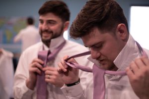 Two men in pink ties and white shirts hold their ties and laugh together at a formal event.