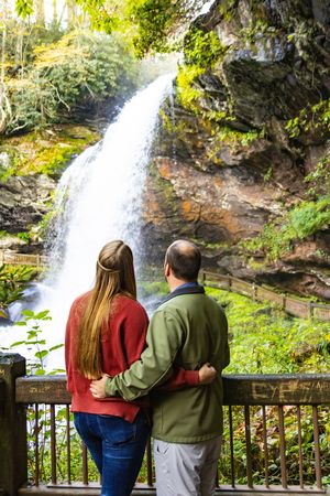 Two people stand at a wooden railing viewing a scenic waterfall in a lush forest setting.