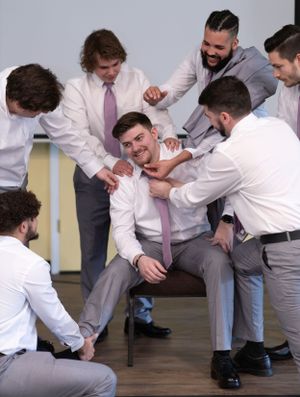 A group of groomsmen in dress shirts and ties playfully interact and laugh together during a candid moment.
