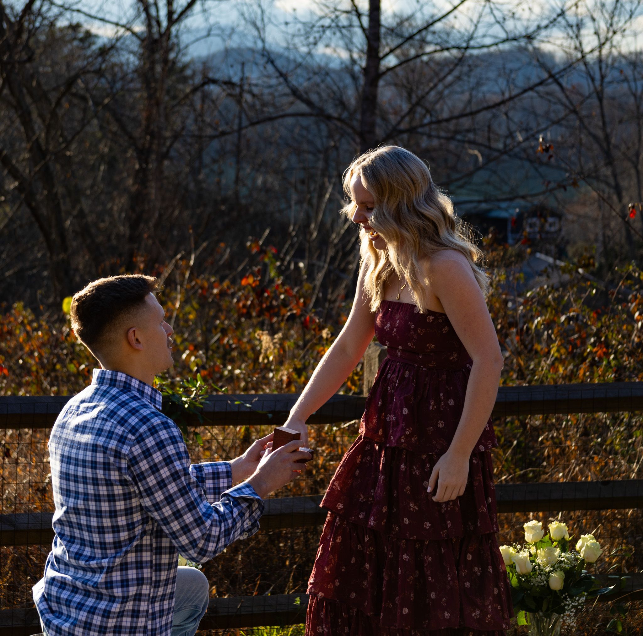 A romantic outdoor proposal scene at sunset with mountains in the background and autumn foliage.