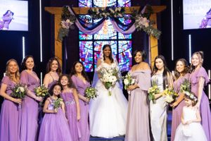 A bridal party in purple bridesmaid dresses and a bride in white stand in front of a stained glass window at a church.