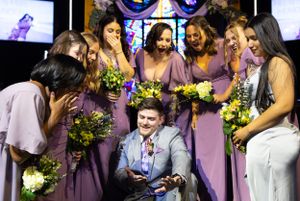 Wedding party in lavender dresses gathers around seated person holding bouquets of yellow flowers in church setting.
