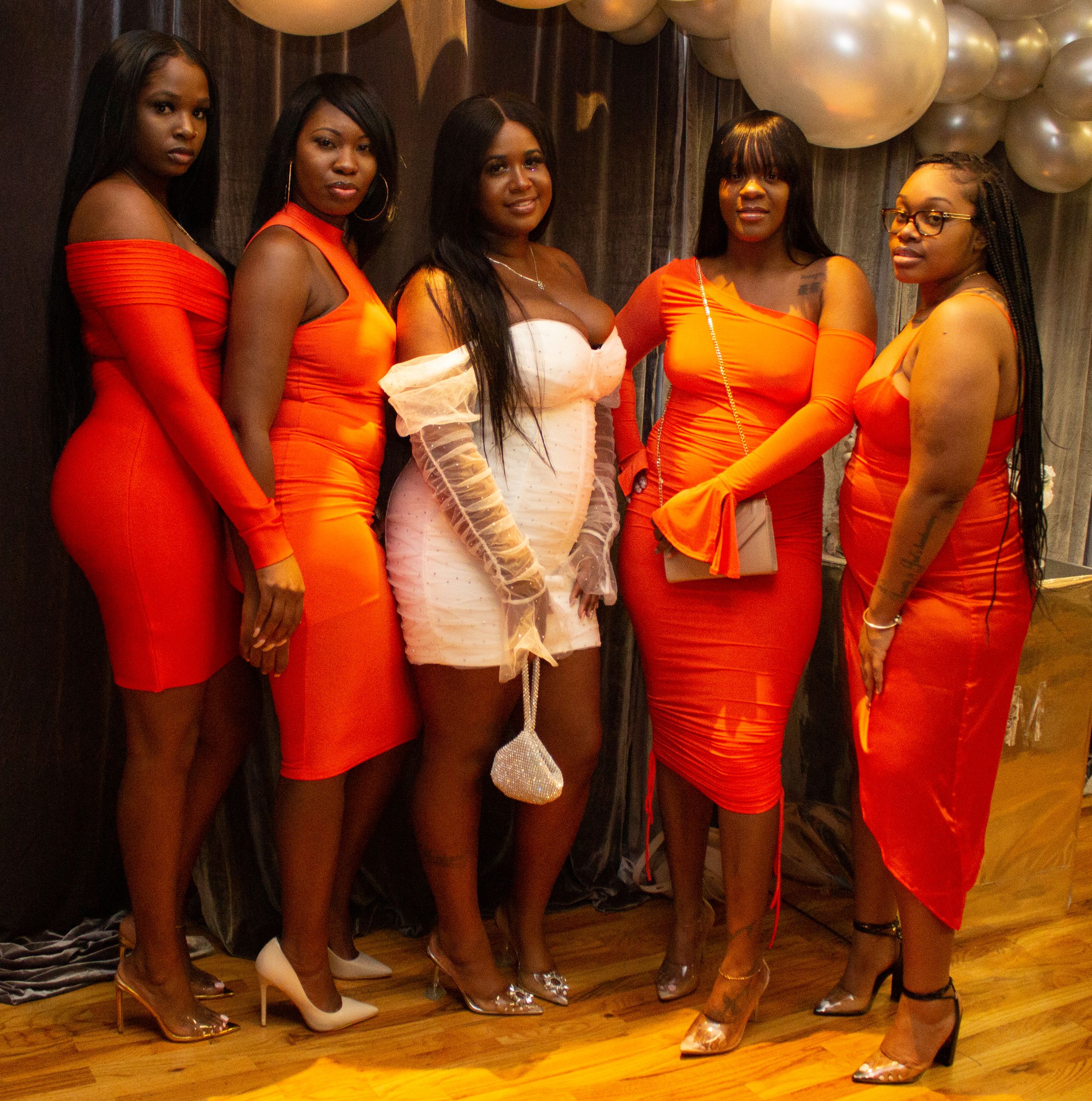 A group of five women in vibrant orange and red fitted dresses pose together at a celebration with balloons in background.