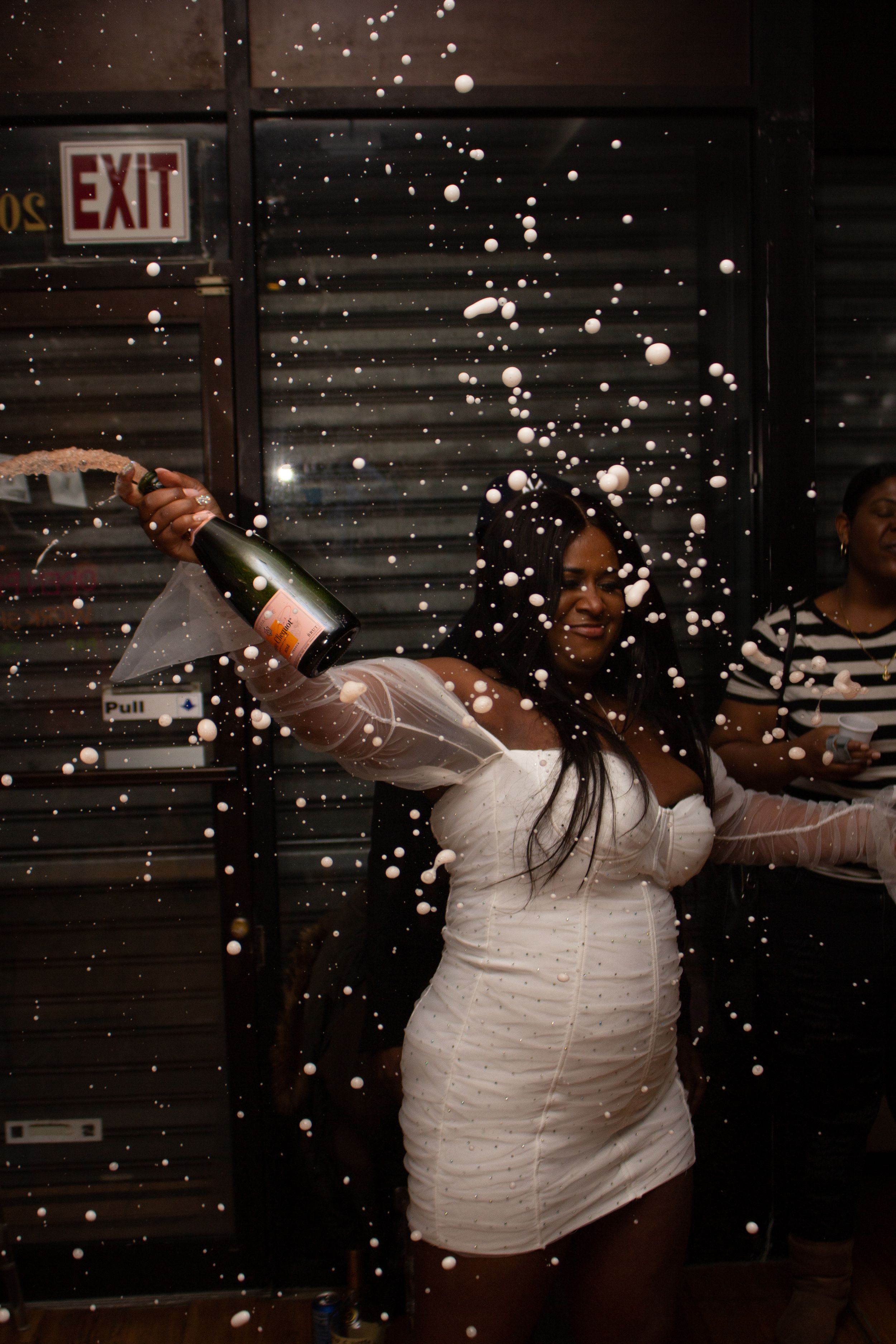 Dancing person in a white dress popping champagne at a party with sparkles and an exit sign in background.