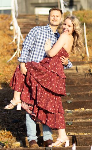A couple poses together on rustic outdoor steps, with her wearing a flowing red polka dot dress and him in plaid.